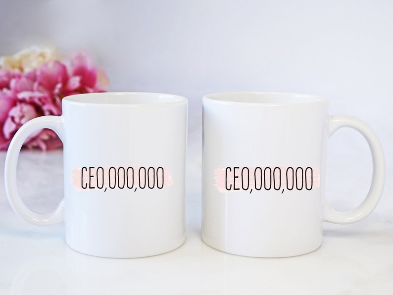 Op de afbeelding: Twee witte keramische mokken met zwarte tekst die "CEO,000,000" op elke mok staat. De tekst staat op een roze aquarel achtergrond.