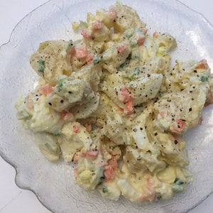May include: A close-up of a white bowl filled with potato salad. The salad is made with potatoes, eggs, celery, carrots, and mayonnaise. The salad is seasoned with black pepper.
