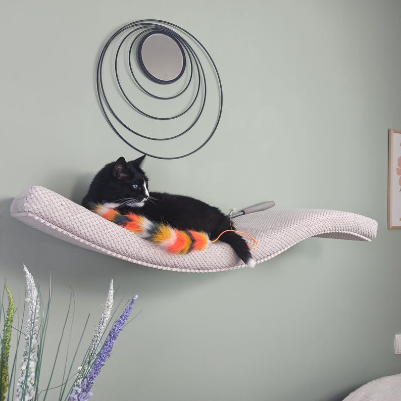 May include: A wall-mounted, curved cat shelf featuring a textured, light beige fabric surface. A black and white cat is resting on the shelf, playing with a long, multi-colored faux fur toy with orange, yellow, and grey stripes.
