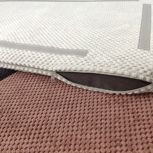 May include: A rectangular cushion cover with a textured cream-colored popcorn knit fabric. The cover features a smooth gray border strip and a side zipper closure with a metal pull. The cushion is placed atop a dark brown and a terracotta textured fabric.