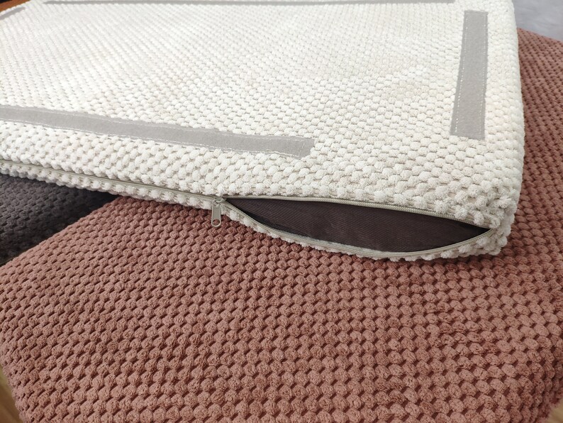 May include: A rectangular cushion cover with a textured cream-colored popcorn knit fabric. The cover features a smooth gray border strip and a side zipper closure with a metal pull. The cushion is placed atop a dark brown and a terracotta textured fabric.