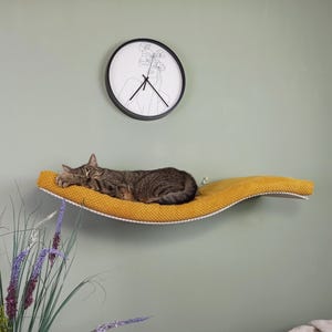 Può includere: Un gatto soriano marrone dorme su una mensola da parete curva e testurizzata color giallo senape. Sopra la mensola è appeso un orologio da parete rotondo con cornice nera che presenta un'illustrazione a linee minimalista di una persona con fiori.