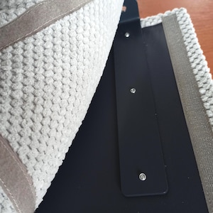 May include: A textured light gray fabric panel with a raised, bumpy surface pattern is partially lifted to reveal a smooth, matte black rectangular base. The black base features three evenly spaced silver metal rivets along its vertical center.
