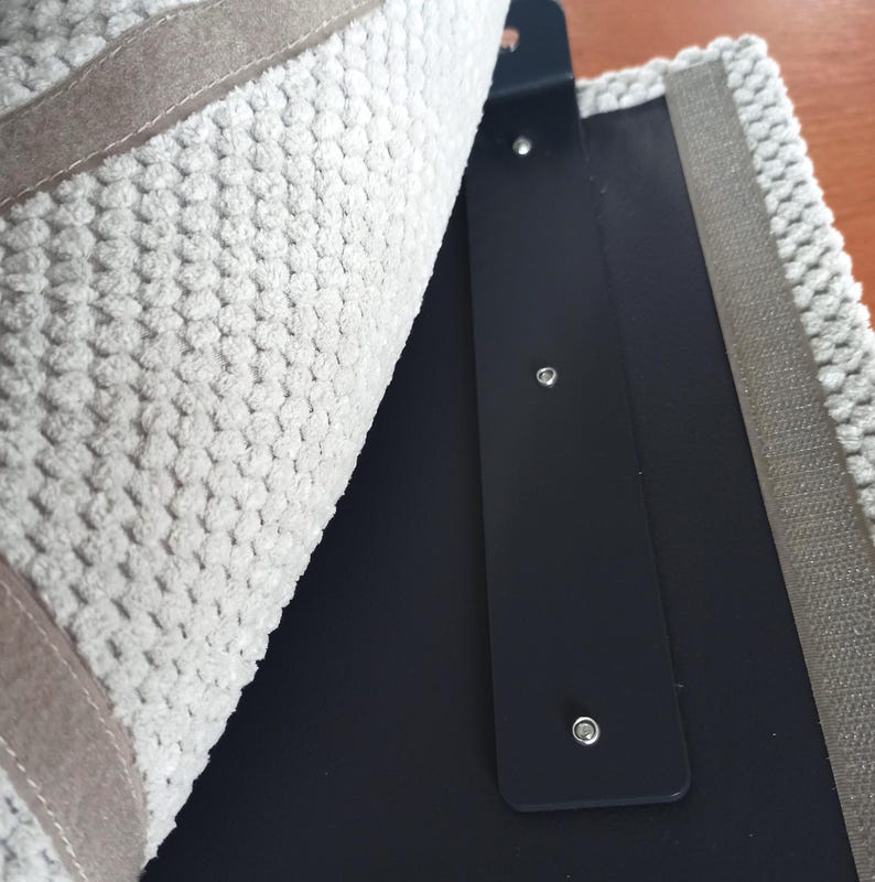 May include: A textured light gray fabric panel with a raised, bumpy surface pattern is partially lifted to reveal a smooth, matte black rectangular base. The black base features three evenly spaced silver metal rivets along its vertical center.