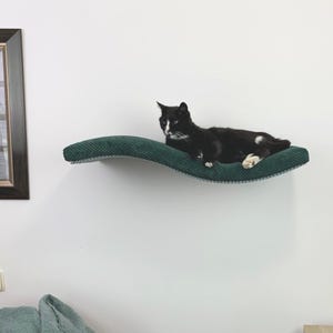 Può includere: Un gatto bianco e nero rilassato su un letto per gatti verde e curvo montato su una parete bianca. Il letto per gatti è realizzato in un materiale morbido e peluche e ha una forma ondulata.
