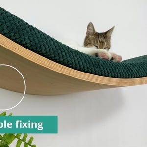 May include: A curved wooden cat bed with a green cushion, mounted to a wall with invisible fixings. A cat is sleeping on the bed.