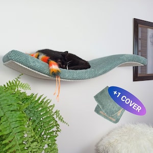 May include: A wall-mounted, curved cat shelf featuring a textured sage green fabric surface. A black cat sleeps on the shelf next to a colorful, multi-striped faux fur toy. An inset graphic shows an additional sage green fabric cover with text +1 COVER.