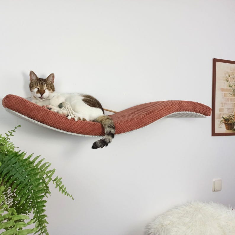 May include: A wall-mounted cat hammock featuring a curved, wave-like design with a textured, rust-colored fabric surface. A white and brown tabby cat is resting on the hammock, which is secured to the wall with visible rope supports.