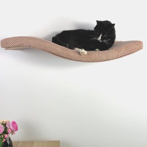 May include: A wall-mounted cat shelf with a curved, wave-like design, upholstered in a textured, light brown fabric. A black and white cat is resting on the surface of the shelf, which is attached to a plain white wall.