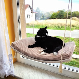 Può includere: Un gatto bianco e nero rilassato su un letto per gatti a forma di amaca beige sospeso da una finestra. Il letto è fatto di tessuto intrecciato e ha una forma curva. Il gatto guarda di lato, con le zampe appoggiate sul letto.