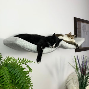 May include: Two cats sleeping on a gray, wave-shaped cat bed mounted on a white wall. The bed is made of soft, plush fabric and has a raised edge for comfort.