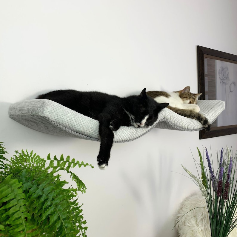 May include: Two cats sleeping on a gray, wave-shaped cat bed mounted on a white wall. The bed is made of soft, plush fabric and has a raised edge for comfort.