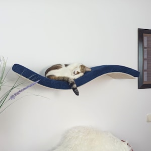 Può includere: Una mensola da parete per gatti dal design curvo a forma di onda, con una superficie superiore in tessuto blu scuro testurizzato e una parte inferiore chiara. Un gatto bianco e marrone è raggomitolato e dorme al centro della piattaforma.