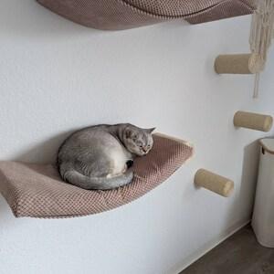 May include: A grey cat curled up on a wall-mounted, brown, textured fabric hammock. To the right, three cylindrical, beige, sisal-wrapped wall steps are mounted vertically, with a decorative macrame element hanging from the top step.