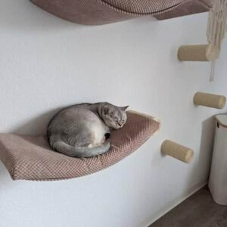 May include: A grey cat curled up on a wall-mounted, brown, textured fabric hammock. To the right, three cylindrical, beige, sisal-wrapped wall steps are mounted vertically, with a decorative macrame element hanging from the top step.