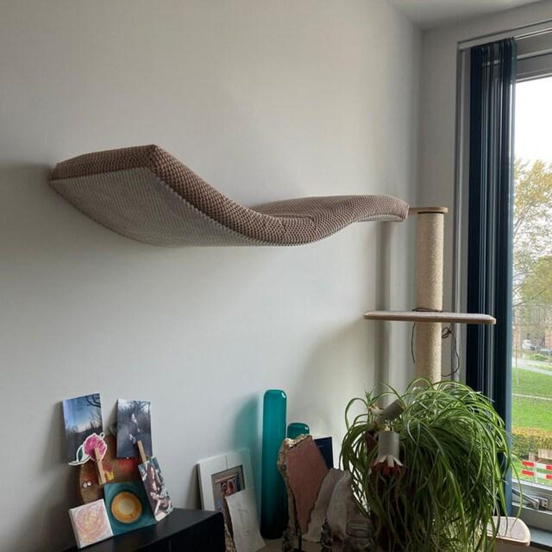 May include: A wall-mounted cat lounge featuring a curved, hammock-style design with a textured, light beige woven fabric surface. The structure is supported by a vertical post wrapped in sisal rope, providing a stable platform for feline rest.