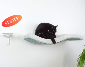 May include: A wall-mounted cat shelf with a curved, textured light gray and teal fabric surface. A black cat is resting on the shelf. To the left, a small white support bracket is visible with an orange oval graphic containing the text +1 STEP.