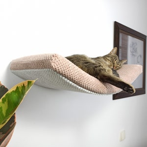 May include: A wall-mounted cat hammock featuring a soft, textured, blush pink fabric top and a light gray underside. The hammock is curved in shape and provides a resting space for a brown tabby cat, which is currently sleeping on the surface.