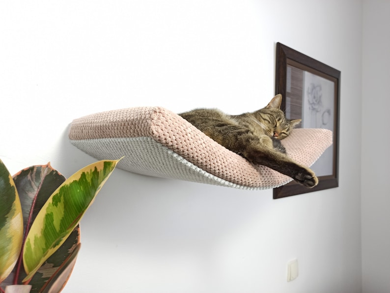 May include: A wall-mounted cat hammock featuring a soft, textured, blush pink fabric top and a light gray underside. The hammock is curved in shape and provides a resting space for a brown tabby cat, which is currently sleeping on the surface.