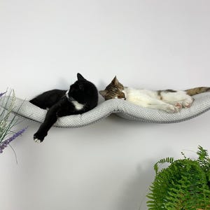 May include: Two cats sleeping on a gray, curved cat bed mounted on a white wall. The cat bed is made of soft, plush fabric and has a wave-like design. The cats are positioned side-by-side, with one cat on each side of the bed.