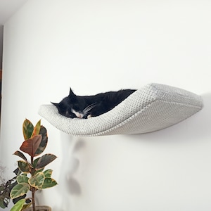 May include: A wall-mounted cat bed featuring a curved, light gray textured fabric surface. The bed is securely attached to a plain white wall, providing a suspended resting area for a black and white cat currently sleeping on the cushioned platform.