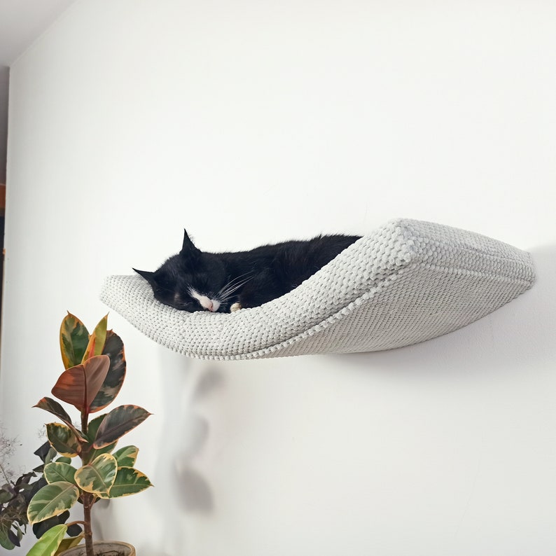 May include: A wall-mounted cat bed featuring a curved, light gray textured fabric surface. The bed is securely attached to a plain white wall, providing a suspended resting area for a black and white cat currently sleeping on the cushioned platform.