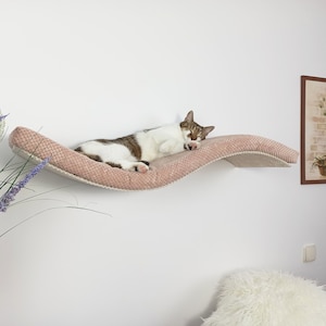 May include: A wall-mounted cat shelf featuring a curved, wave-like design with a textured, dusty pink fabric surface. The shelf is mounted horizontally and supports a sleeping white and brown tabby cat resting along its length.