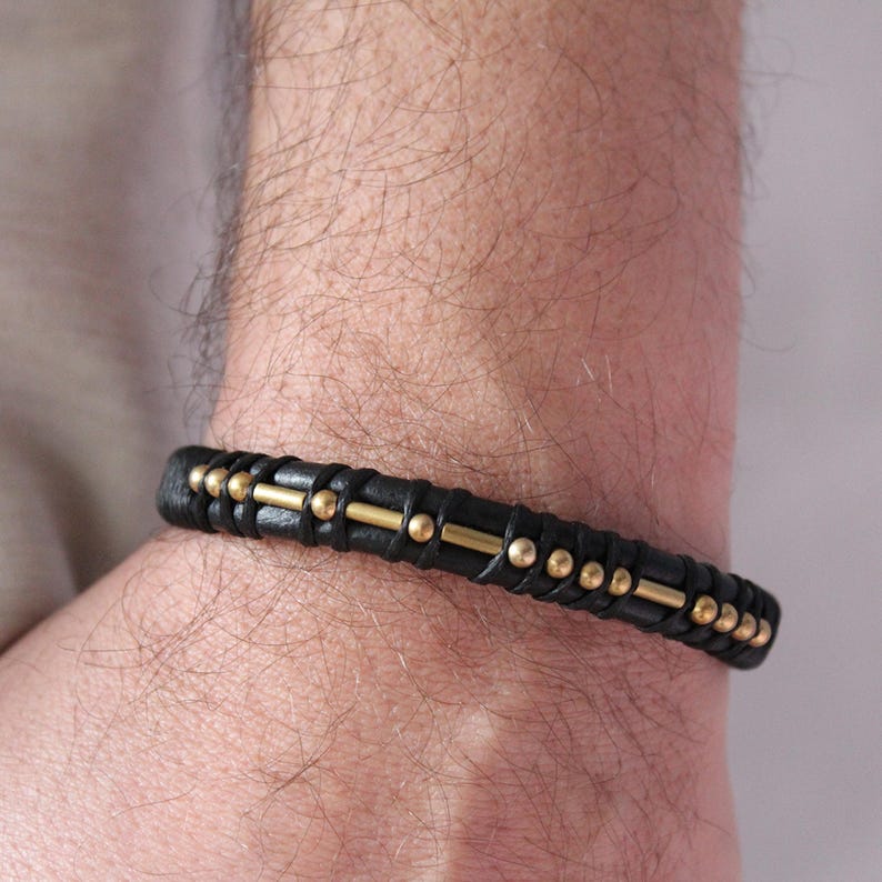 May include: A black leather bracelet with gold accents. The bracelet has a braided design and is adorned with small gold beads.