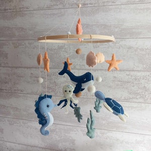 Op de afbeelding: Een baby mobile met vilten zeedieren, waaronder een walvis, zeepaardje, octopus en schildpad. De mobile heeft een houten ring en is versierd met oranje, blauwe en beige vilten elementen. Witte vilten ballen en zeesterren dragen bij aan het oceaanthema.