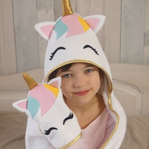 Può includere: Asciugamano con cappuccio bianco con un design a forma di unicorno. Il cappuccio ha un corno dorato, orecchie rosa e blu e occhi ricamati. L'asciugamano ha un bordo dorato e un asciugamano per le mani a forma di unicorno abbinato.