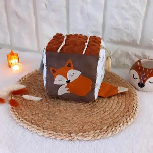 Könnte beinhalten: Ein brauner und orangefarbener Stoffwürfel mit einem schlafenden Fuchs-Design. Der Würfel hat verschiedene Texturen und einen daran befestigten Holzring.