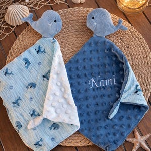 Könnte beinhalten: Zwei weiche Baby-Schmusetücher mit Plüsch-Wal-Köpfen. Ein Tuch hat einen hellblauen Stoff mit dunkelblauem Walmuster und eine weiße genoppte Unterseite. Das andere ist dunkelblau, hat eine genoppte Textur und den eingestickten Namen Nami.
