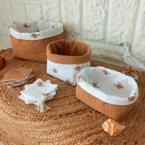 Peut inclure: Trois paniers de rangement en tissu de différentes tailles, avec un extérieur marron et un intérieur blanc orné de motifs de baleines oranges. Plusieurs débarbouillettes assorties sont également visibles, ainsi qu'un coquillage et une figurine d'oiseau décorative.
