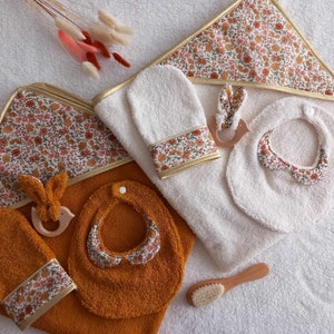 Op de afbeelding: Twee badcapes met bijpassende slabben en wanten. Eén cape is wit met een bloemenpatroon en de andere is mosterdgeel met een bloemenpatroon. Beide capes hebben een gouden rand. De slabben en wanten zijn gemaakt van een zacht, absorberend materiaal.