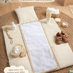 Peut inclure: Tapis à langer nomade de couleur crème avec un centre blanc et une bordure beige. Il est accompagné de produits de soins pour bébés, d'un jouet en bois et de petites chaussures marron. Le texte indique "TAPIS DE CHANGE NOMADE" et "PRODUIT ARTISANAL".