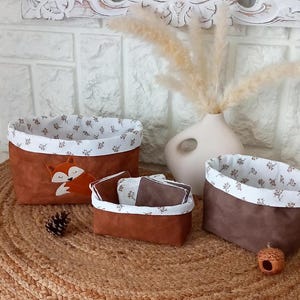 Peut inclure: Trois paniers de rangement en tissu marron avec une doublure en tissu blanc et un motif de renard sur le plus grand panier. Les paniers sont sur une surface en osier marron.