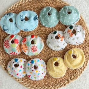 Pode incluir: Uma coleção de babadores para bebês com vários padrões, incluindo baleias, estrelas, flores, raposas e formas geométricas. Os babadores são em tons de azul, verde, laranja, branco e amarelo, dispostos em uma esteira tecida.