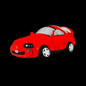Puede incluir: Un coche de juguete de peluche rojo con forma de coche deportivo. El coche tiene faros negros y un interior blanco.