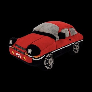 Op de afbeelding: Rode pluche speelgoedauto met zwarte accenten en grijs stof op het dak. De auto heeft een gedetailleerde voorkant met koplampen en een grille.