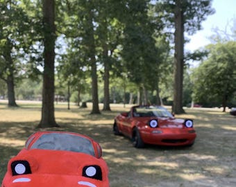 May include: A red plush toy car with white eyes and a black mouth. The toy car is sitting on a wooden stump in a grassy area. A red convertible car is in the background.