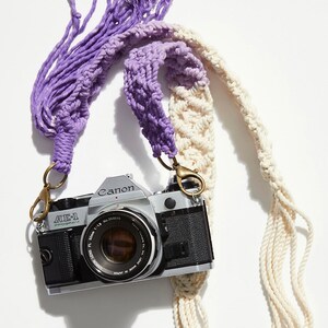 May include: A vintage Canon AE-1 camera with a purple and white crocheted camera strap. The camera has a silver body and a black lens. The strap has long tassels on both ends.