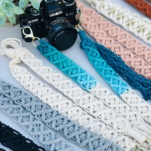 May include: A set of seven camera straps made of macrame with different color combinations. The straps are made with a white, gray, blue, and black color scheme. The straps have a metal clasp on one end and a tassel on the other end.