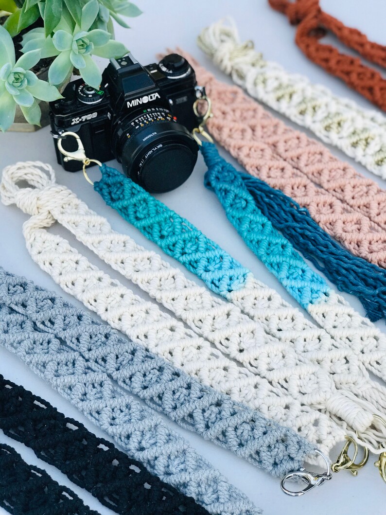 May include: A set of seven camera straps made of macrame with different color combinations. The straps are made with a white, gray, blue, and black color scheme. The straps have a metal clasp on one end and a tassel on the other end.