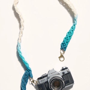 May include: A white and blue ombre camera strap with a tassel. The strap is made of macrame and has a gold metal clasp. A silver Canon AE-1 camera is attached to the strap.