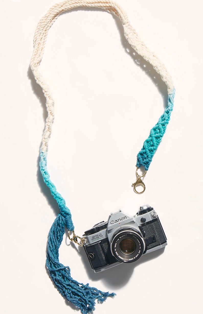 May include: A white and blue ombre camera strap with a tassel. The strap is made of macrame and has a gold metal clasp. A silver Canon AE-1 camera is attached to the strap.