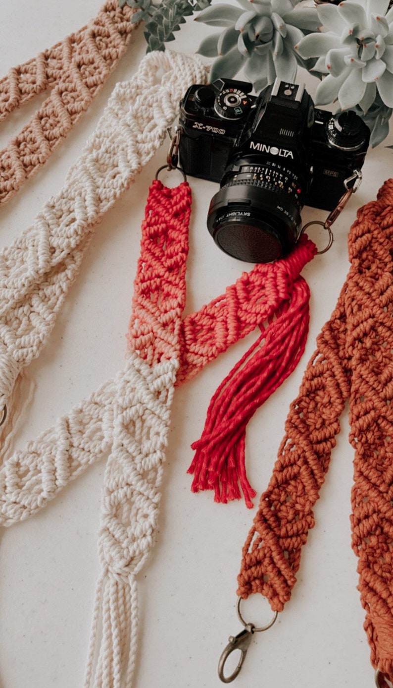 May include: Three camera straps made of macrame with different color combinations. The straps are made of cotton cord and have a tassel at the end. The straps are shown with a black camera with a MINOLTA lens.