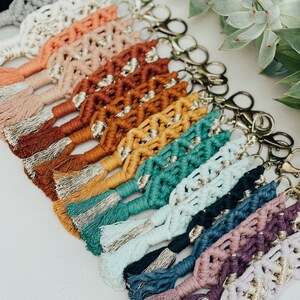 May include: A collection of colorful macrame keychains with gold accents and tassels. The keychains are arranged in a row, with the tassels hanging down. The keychains are made with a variety of colors, including red, orange, yellow, green, blue, purple, and pink.