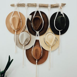 May include: A wall-mounted hat rack made of driftwood and macrame rope with six hats hanging from it. The hats are in various colors and styles, including brown, beige, and green.