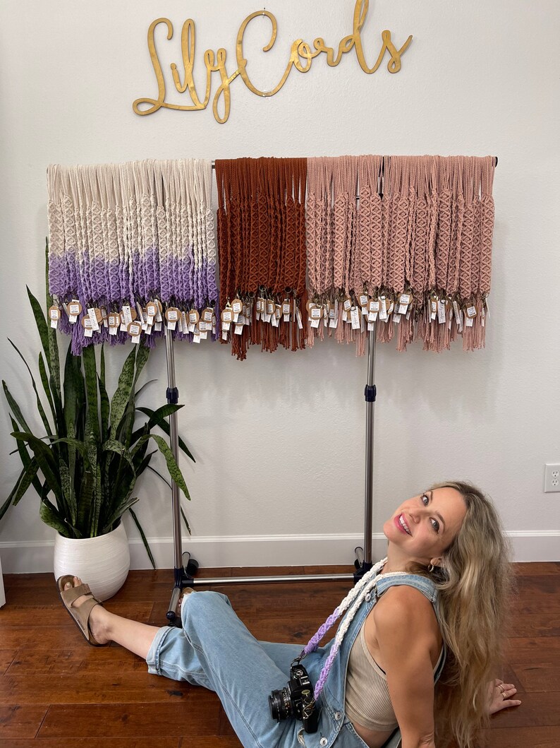 May include: A woman sits in front of a rack of handmade macrame keychains. The keychains are in three colors: white with purple, brown, and pink. The rack is labeled "Lily Cords".