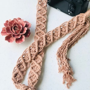May include: A light brown camera strap made of macrame with a tassel at the end. The strap is lying on a white surface with a ceramic flower and a black camera.